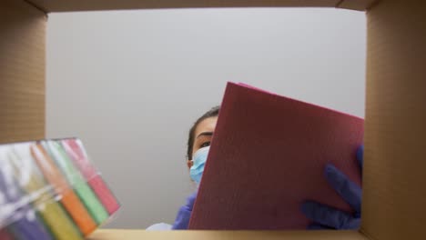 Woman-in-Mask-Taking-Cleaning-Supplies-From-Box.home-delivery,-shipping-and-pandemic-concept-woman-in-protective-medical-mask-and-gloves-opening-parcel-box,-looking-inside-and-taking-cleaning-supplies-out